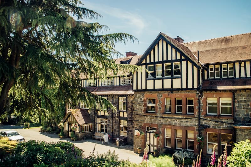 The charming facade of Lake Vyrnwy Hotel, a historic country house with Tudor-style black-and-white timber detailing and stone walls. The building is surrounded by lush greenery, with a large tree framing the image and a well-manicured garden in the foreground. A bright blue sky and dappled sunlight enhance the inviting atmosphere.
