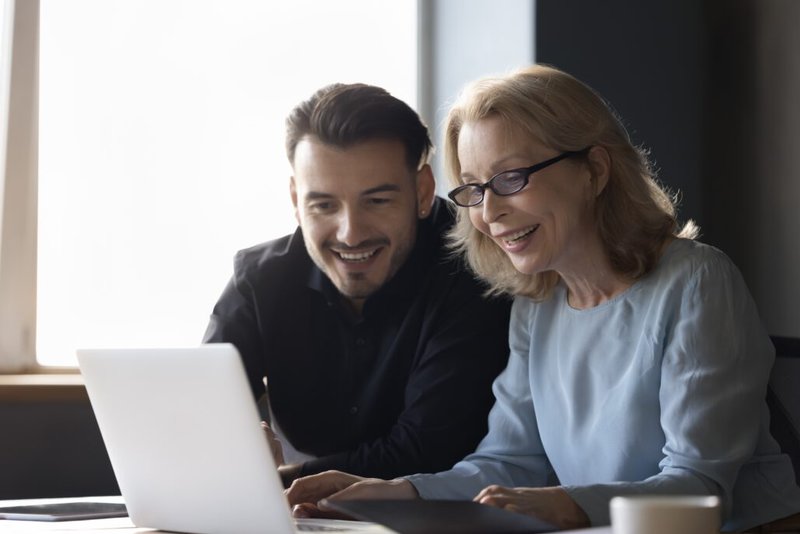 Ein Mann und eine Frau, beide Kolleg*innen, sitzen nebeneinander und blicken gut gelaunt auf einen Laptop. Die positive Stimmung zeigt, wie wertschätzende Kommunikation Zusammenarbeit stärkt und ein angenehmes Miteinander im Team fördert.