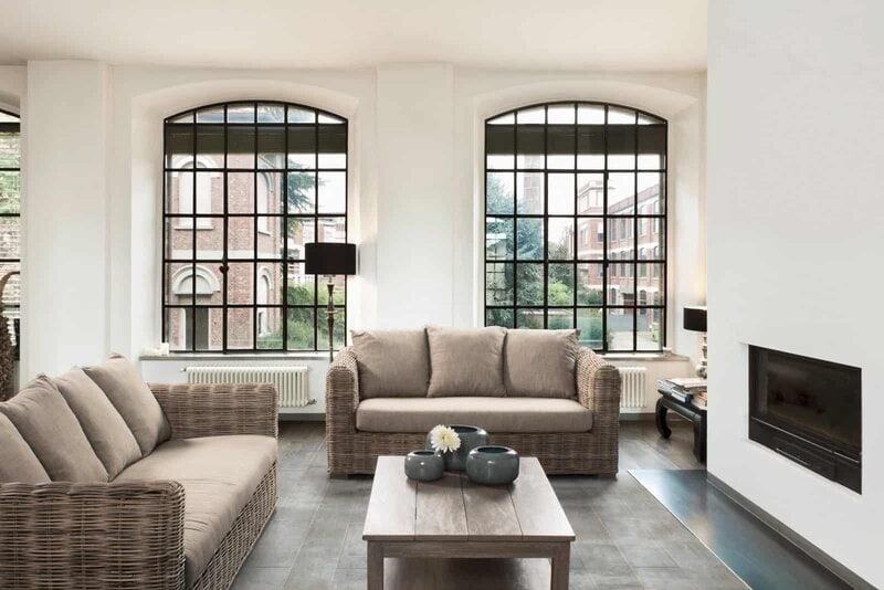 Soggiorno luminoso con grandi finestre in ferro nero, arredi in rattan e comfort moderno.