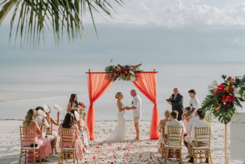 Amanda and Rob say their vows under a coral-red arch on the beach at Baan Puri, Koh Samui, with guests on gold chairs