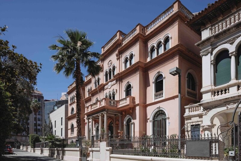 Elegantes historisches Gebäude mit mediterraner Architektur, verzierten Fassaden und Palmen in einer sonnigen Stadt.