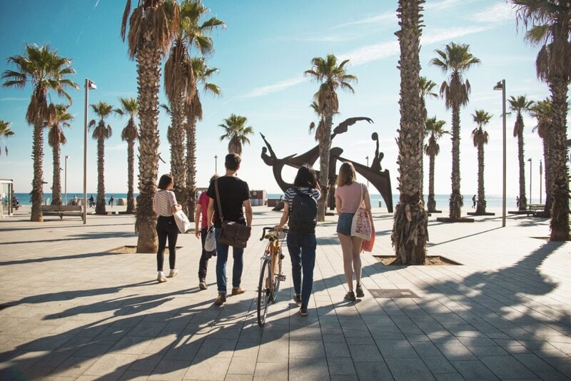 Eine Gruppe junger Menschen spaziert entlang einer sonnigen Palmenpromenade am Meer in Richtung einer modernen Skulptur.