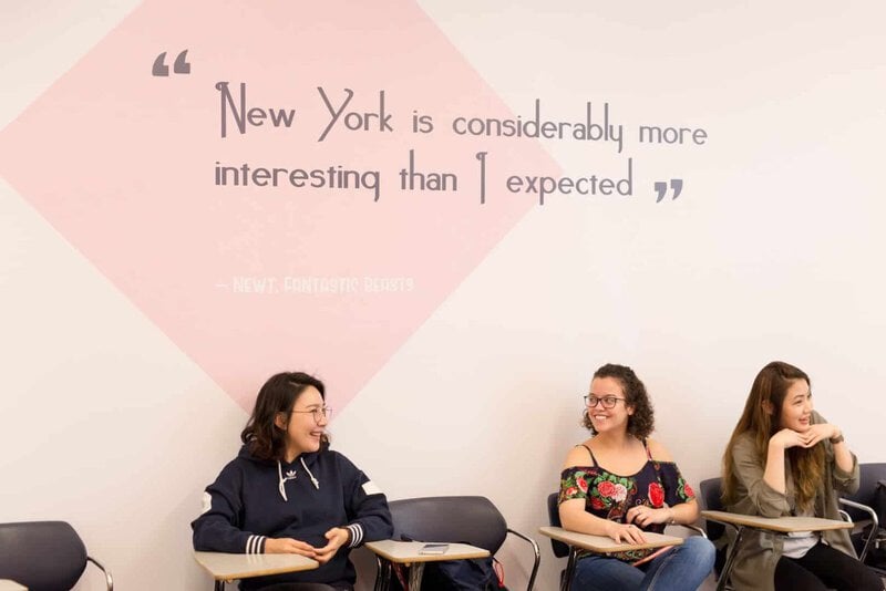 Drei junge Frauen sitzen in einem Klassenzimmer und unterhalten sich lachend vor einer Wand mit einem inspirierenden Zitat über New York.