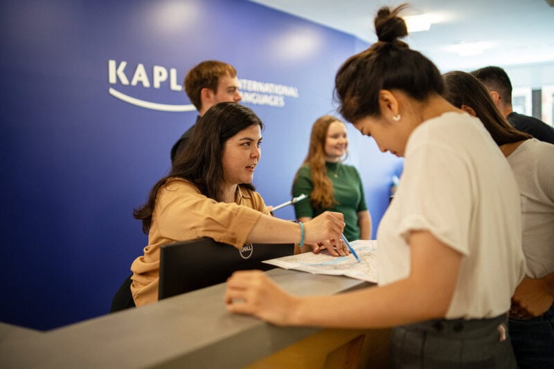 Eine Mitarbeiterin am Empfang einer Sprachschule erklärt einer Studentin etwas auf einer Karte, während andere Personen im Hintergrund warten.