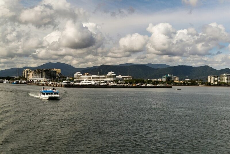 Hafenstadt mit modernen Gebäuden, Yachten und einem Ausflugsboot vor einer beeindruckenden Bergkulisse unter bewölktem Himmel.