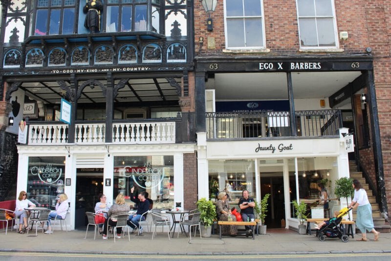 Cafés und Geschäfte in Chester mit historischem Fachwerkgebäude und Menschen, die draußen sitzen und das Stadtleben genießen.