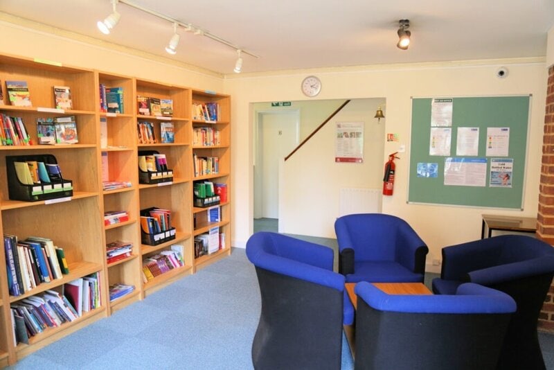 Gemütlicher Aufenthaltsraum mit Bücherregalen, blauen Sesseln und einer Infotafel in einer Sprachschule oder Bildungseinrichtung.
