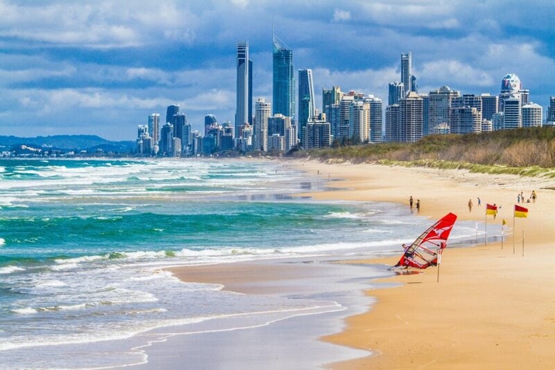 Goldener Sandstrand mit Wellen, Rettungsflaggen und einem Windsurfer, vor der Skyline einer modernen Küstenstadt.
