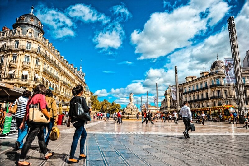 Der Place de la Comédie in Montpellier mit der Fontaine des Trois Grâces, historischen Gebäuden und lebhaftem Stadtleben.