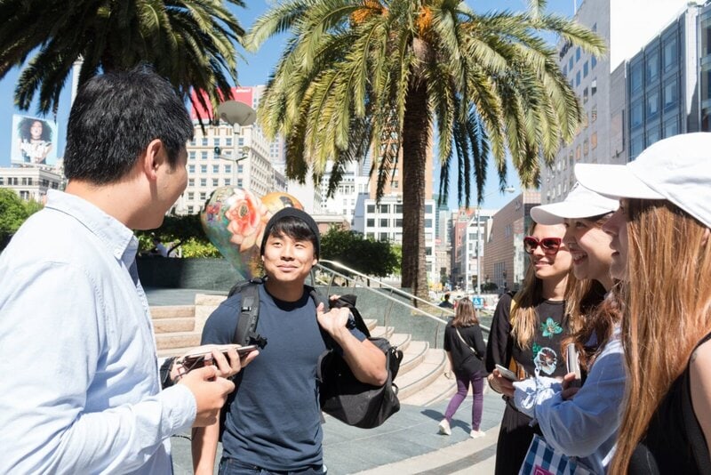 Eine Gruppe junger Menschen unterhält sich fröhlich im Union Square in San Francisco, umgeben von Palmen und moderner Architektur.