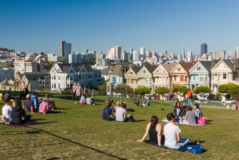 Menschen entspannen im Alamo Square Park mit Blick auf die Painted Ladies und die Skyline von San Francisco.