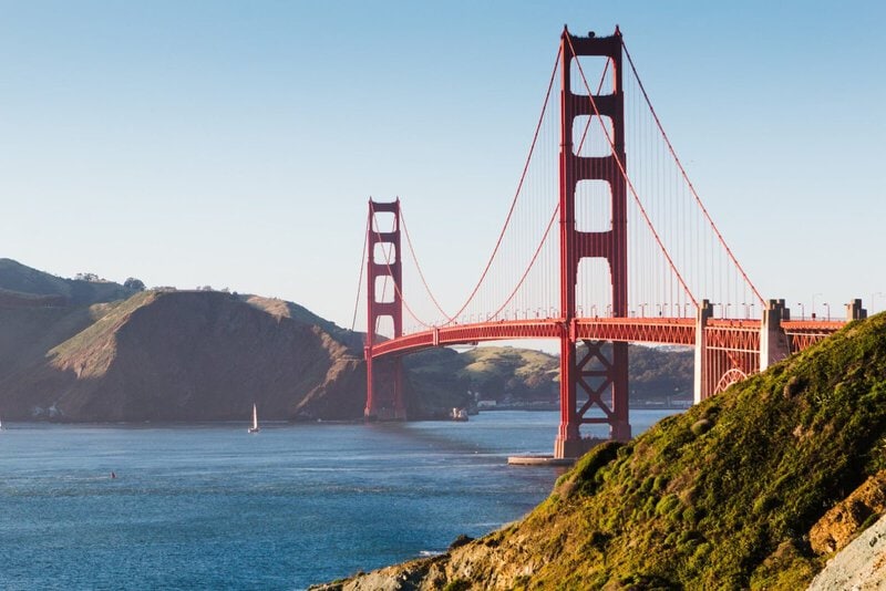Die Golden Gate Bridge in San Francisco an einem sonnigen Tag mit klarem Himmel, Blick auf das blaue Wasser und grüne Hügel im Vordergrund.