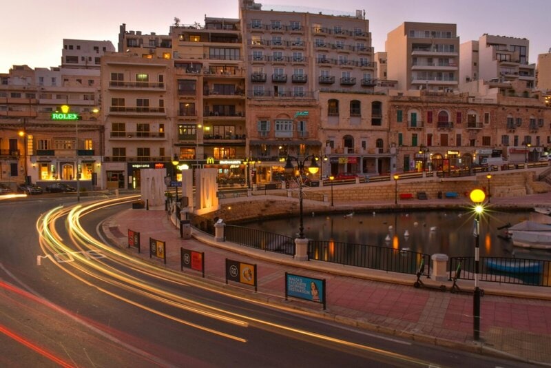 St. Julian’s Bay in Malta bei Nacht mit leuchtenden Lichtspuren des Verkehrs und beleuchteten Gebäuden.