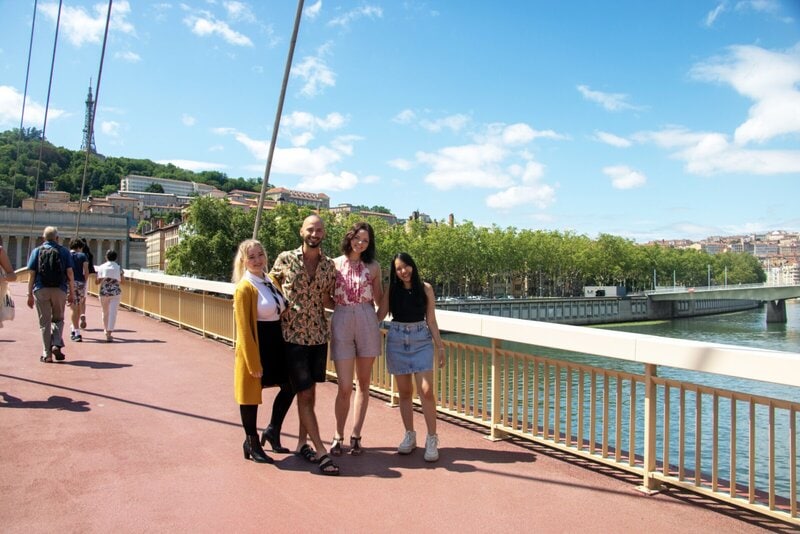 Eine Gruppe junger Menschen auf einer Brücke mit Blick auf die Stadtlandschaft und einen Fluss bei sonnigem Wetter.