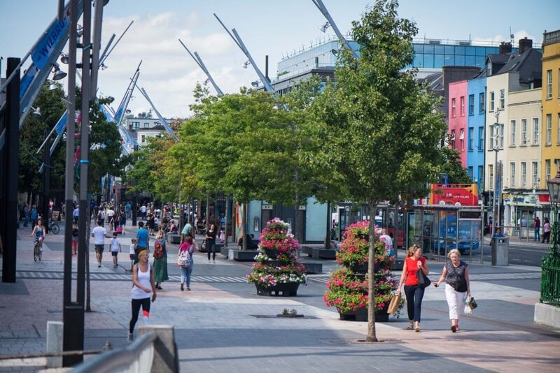 Belebte Einkaufsstraße in Cork, Irland, mit modernen Skulpturen, bunten Gebäuden und Fußgängern.
