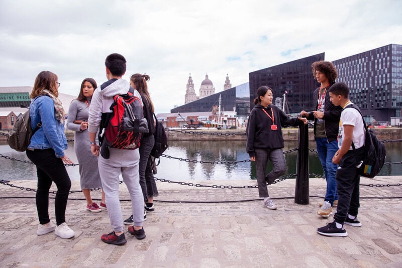Internationale Studierende genießen eine Stadttour am Liverpool Waterfront mit Blick auf moderne Architektur und historische Wahrzeichen.