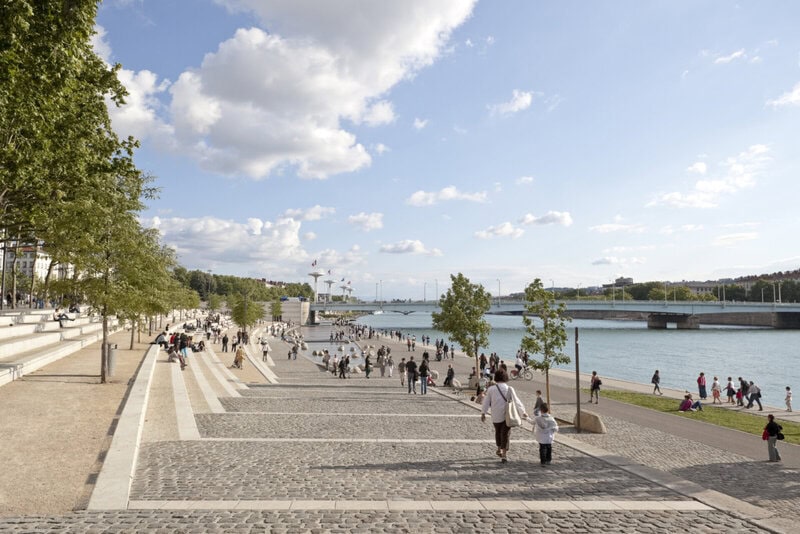 Belebte Uferpromenade mit breiten Treppenstufen, Spaziergängern und Blick auf den Fluss und eine moderne Brücke.