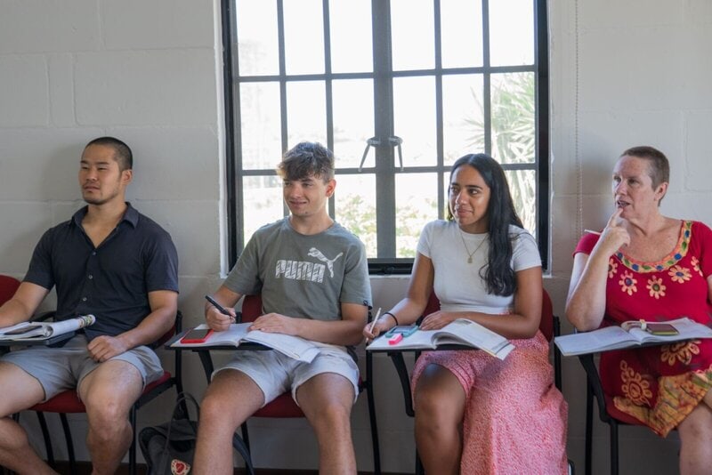 Vier internationale Sprachschüler sitzen in einem Klassenzimmer, nehmen Notizen und hören aufmerksam zu.
