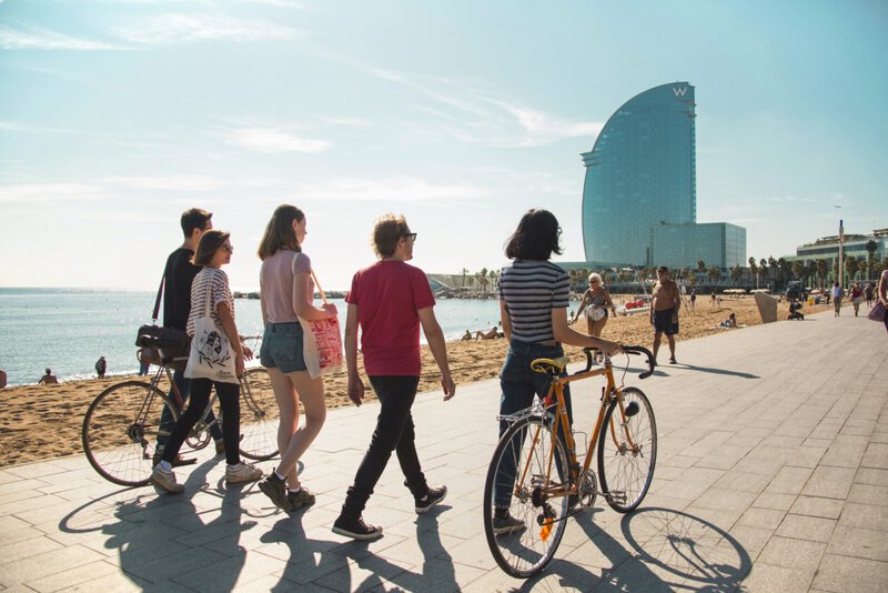 Gruppe junger Menschen spaziert mit Fahrrädern entlang der Strandpromenade in Barcelona mit Blick auf das W-Hotel.