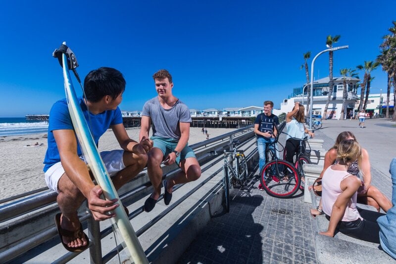 Junge Menschen genießen die Sonne und unterhalten sich an einer Strandpromenade mit Surfbrett und Fahrrädern.