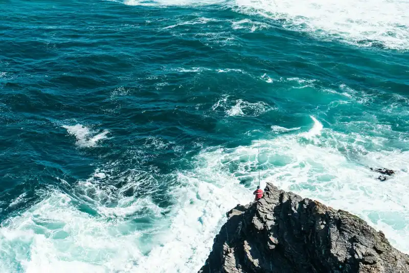 Angler in a red jacket fishing from jagged coastal rocks above deep turquoise ocean surf.