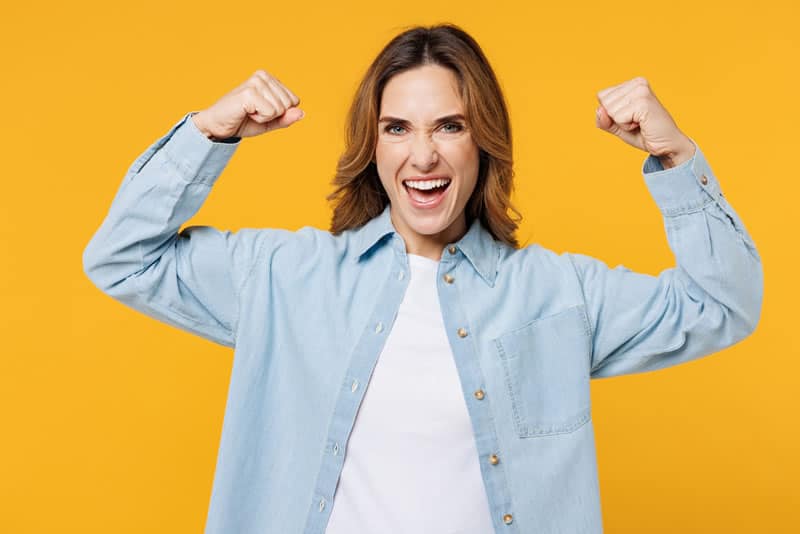 Excited woman in a light blue shirt with arms raised in celebration, smiling and standing against a bright yellow background. Happy she mastered grant reporting