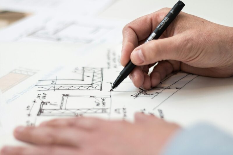 hand holds a pen, drawing on architectural plans spread on a surface.