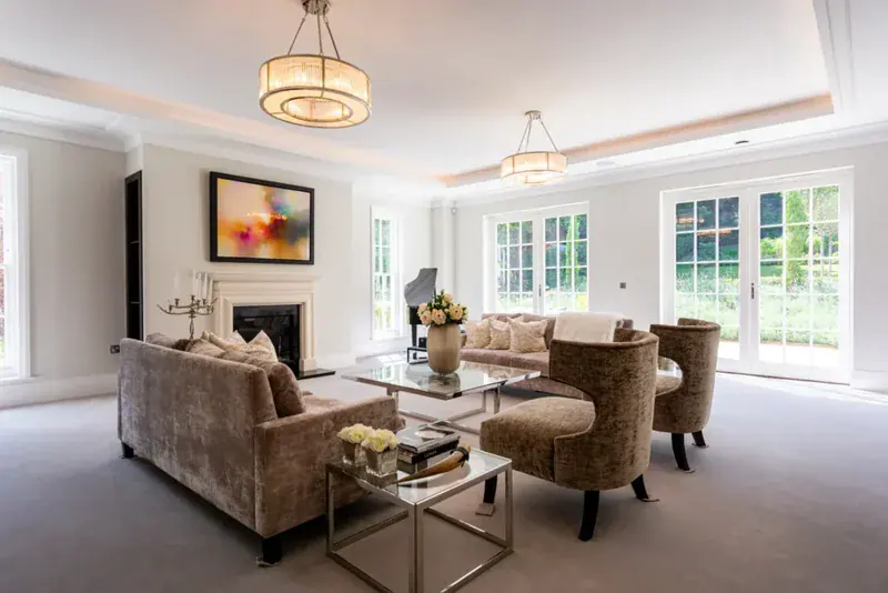 Modern, bright living room with plush taupe sofas, glass coffee tables, large windows, home automation. A fireplace with abstract art above, two chandeliers, and a piano in the corner overlook a green garden in Cobham Surrey.