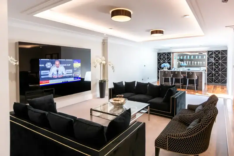 Modern living room with black velvet sofas, a glass coffee table, and a home cinema with smart lighting installation. Ceiling lights illuminate the sleek space, whilst a stylish bar area with glass shelves and wine racks adds elegance in Cobham, Surrey.