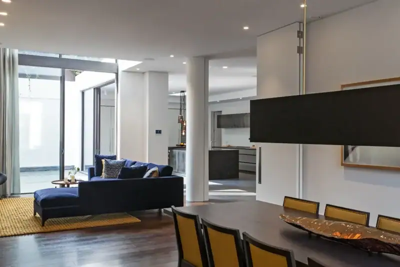 Modern open-plan living and dining area with dark wood floors, a navy blue sofa, yellow rug, large windows, contemporary kitchen, and sleek home automation installation in Hampstead for ultimate convenience.