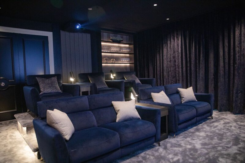 A luxurious home theatre room, showcasing a luxury home cinema installation in Walton-on-Thames, features tiered navy blue sofas, grey cushions, side tables, dim lighting, plush carpet, and shelves displaying glassware against a dark wall.
