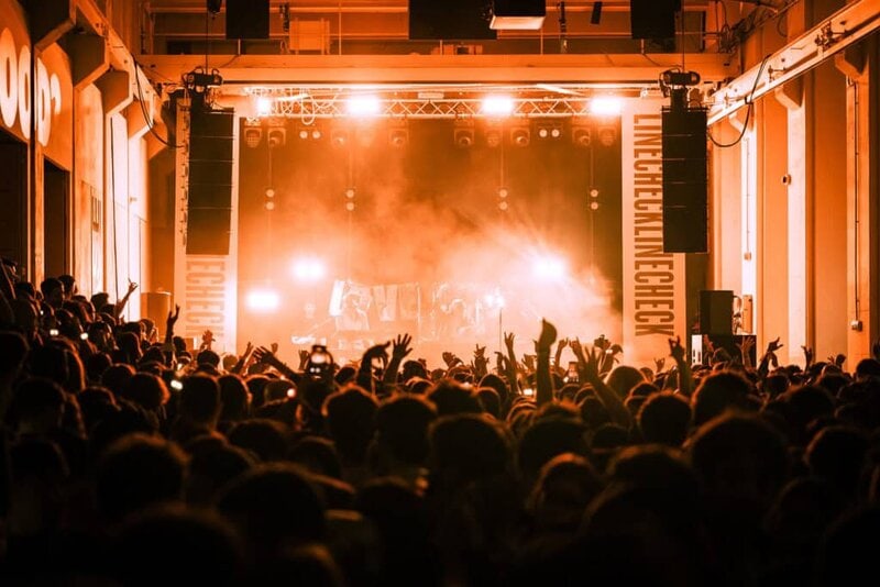 A large crowd in front of the Linecheck stage, illuminated by warm orange lights, with people raising their hands during a live concert.