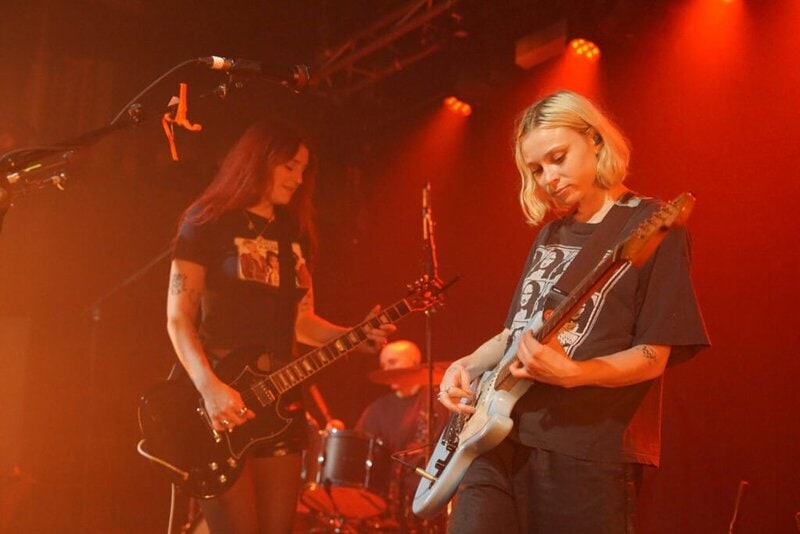 Two musicians from the Momma band play electric guitars on stage under orange lights, with a drummer in the background. Both guitarists wear black T-shirts and display their tattoos, creating an energetic and focused atmosphere.
