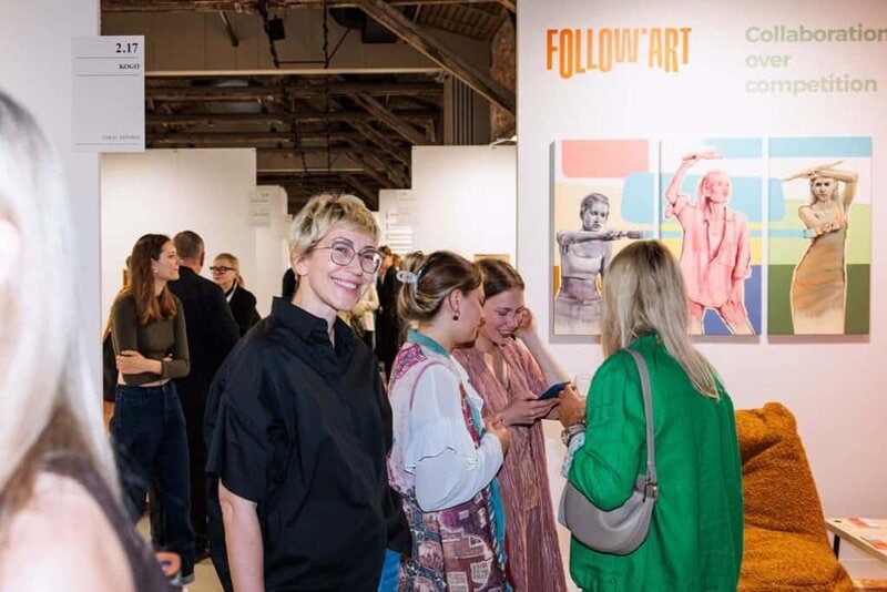 A group of people socialize and smile at an art gallery event. Colorful artwork and a Collaboration over competition sign are visible in the background, highlighting FOLLOW.ART: A New Professional Tool for Artists and Curators. One woman in glasses smiles at the camera.
