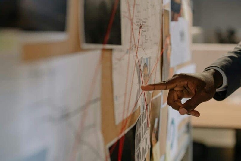 Website portfolio 3 Close-up of an investigator pointing at an evidence board with connected clues using red strings.