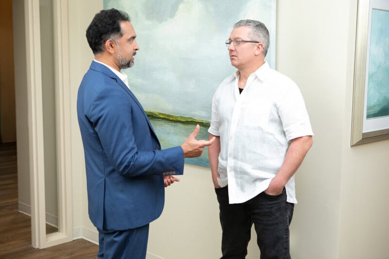 Two men stand and talk in front of a landscape painting; one, Dr. Murthy, wears a gray suit, while the other sports a dark casual outfit.