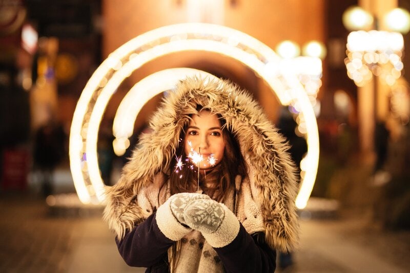 A brown-haired woman in a thick coat holds a sparkler and smiles.