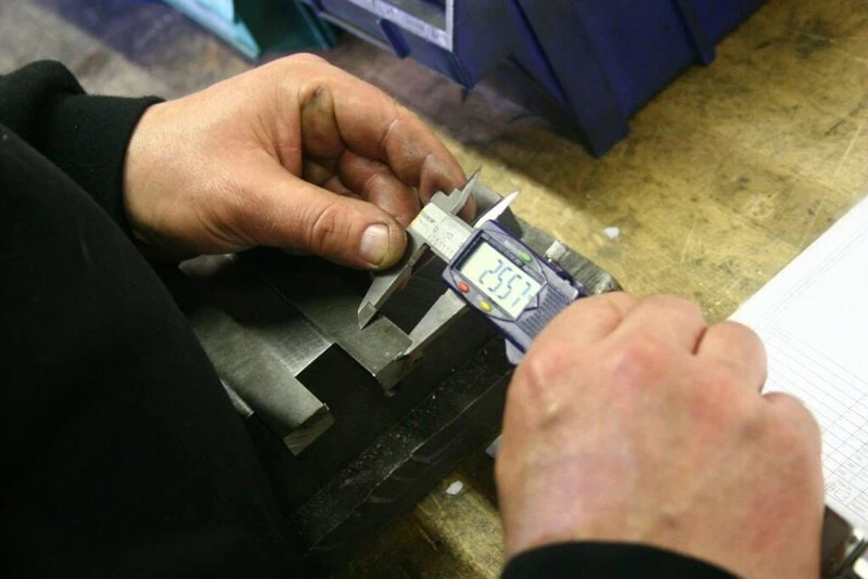 Hands using a digital caliper on a machined metal block; stock illustration of dimensional inspection, not a HecosTech inspection record.