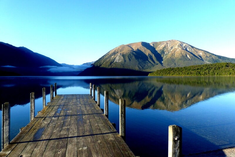 Ein hölzerner Steg ragt in den spiegelglatten Lake Rotoiti im Nelson Lakes Nationalpark, Neuseeland, mit beeindruckenden Bergen im Hintergrund.