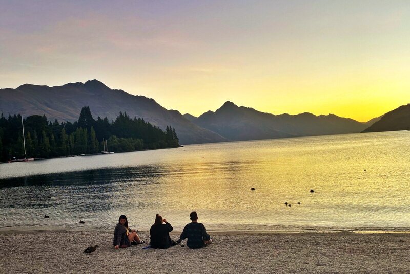 Drei Personen sitzen am Ufer des Lake Wakatipu in Queenstown, Neuseeland, und genießen den goldenen Sonnenuntergang mit Bergpanorama.