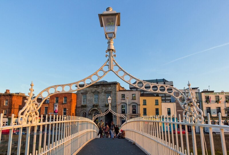 Die historische Ha’penny Bridge in Dublin bei Sonnenuntergang mit Blick auf die farbenfrohen Gebäude am Flussufer.