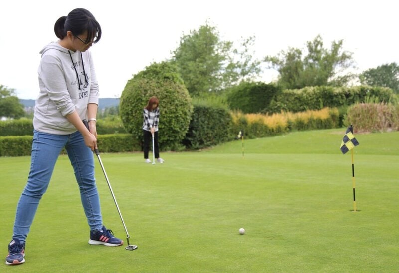 Zwei Frauen spielen Golf auf einem gepflegten Putting Green in einer grünen Parklandschaft.