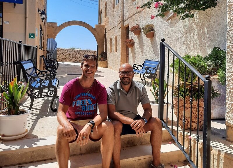 Zwei Männer sitzen entspannt auf einer Treppe in einer malerischen, mediterranen Gasse mit Steinmauern, Blumen und einem historischen Torbogen im Hintergrund.