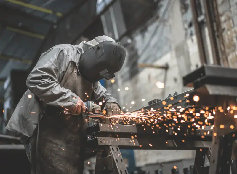 Arbeiter mit Schutzhelm und Schweißermaske schleift Metall in einer Werkstatt – Funken sprühen während der Metallbearbeitung. Symbolbild für Arbeitssicherheit, Staub- und Partikelexposition in der Metallindustrie.