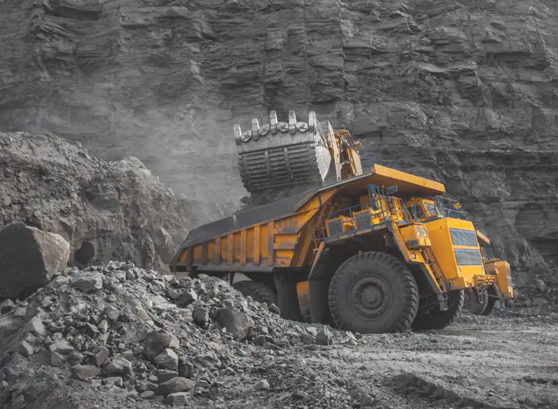 Großer Muldenkipper wird in einem Steinbruch von einem Bagger mit Gestein beladen – typische Szene aus dem Bergbau mit Staubentwicklung und schwerem Gerät.