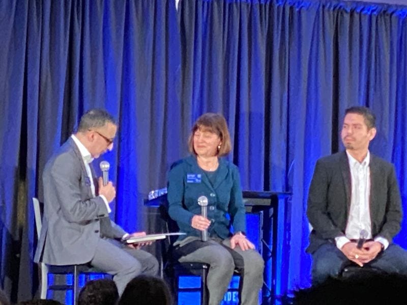 Three individuals on stage in a panel discussion with microphones, seated in front of a blue curtain