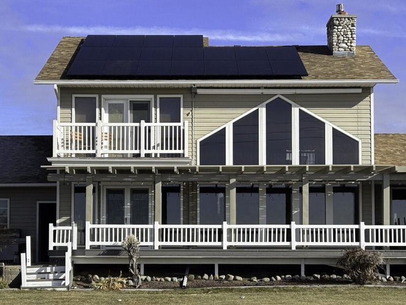 View of the back of a home with a roof-mounted solar array, several windows, a deck, and a grassy yard in the foreground