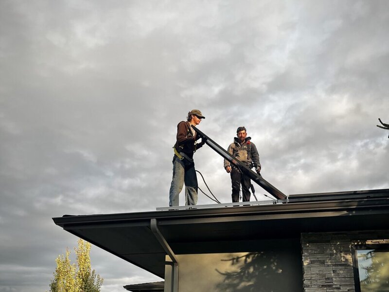 Two individuals on a roof, and one is holding a solar panel. The sky is cloudy in the background.
