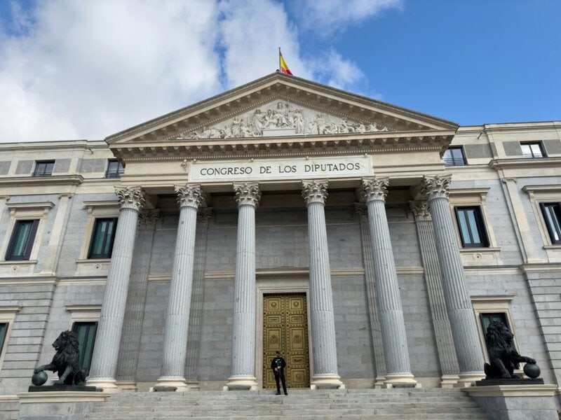 Fachada del Congreso de los Diputados con columnas y puerta dorada, flanqueada por estatuas de leones. Un guardia se encuentr