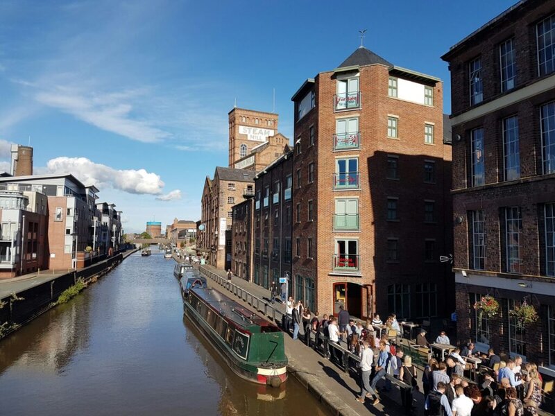 Historischer Kanal in Chester mit schmalen Booten, roten Backsteingebäuden und einem belebten Uferbereich mit Menschen.